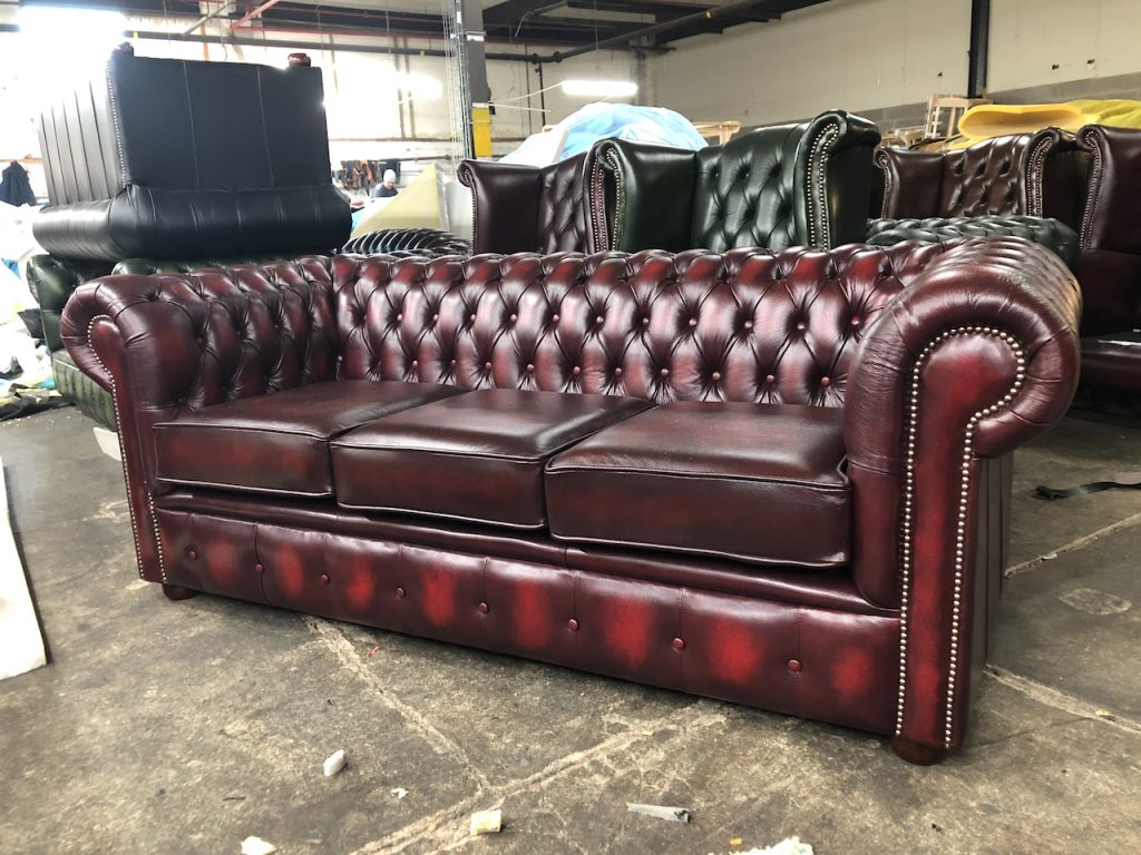 Classic burgundy Chesterfield sofa with deep button tufting, rolled arms, and nailhead trim, placed in a warehouse setting among other furniture pieces.
